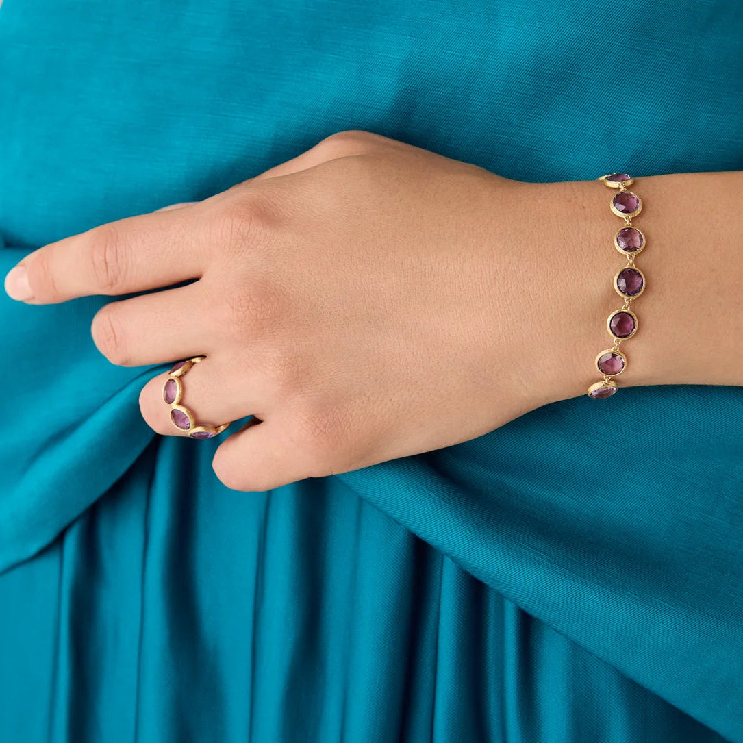 A close-up of a person's hand and wrist wearing a gold bracelet with amethyst gemstones, paired with a matching gold ring on the finger, set against a teal draped fabric background.