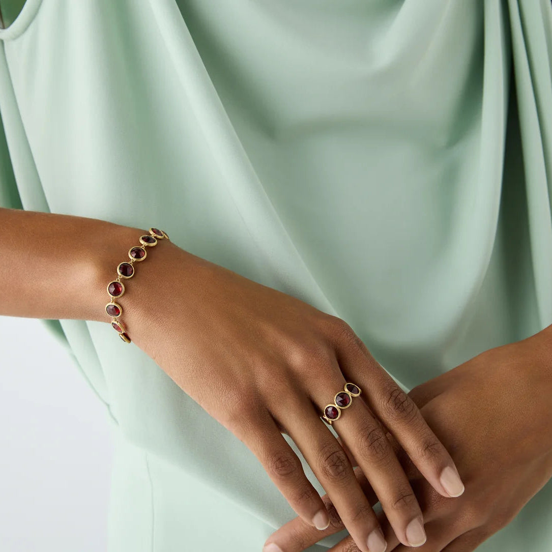 A close-up of a person wearing a gold bracelet and matching ring, each featuring oval red gemstone accents, styled with a mint green draped top.