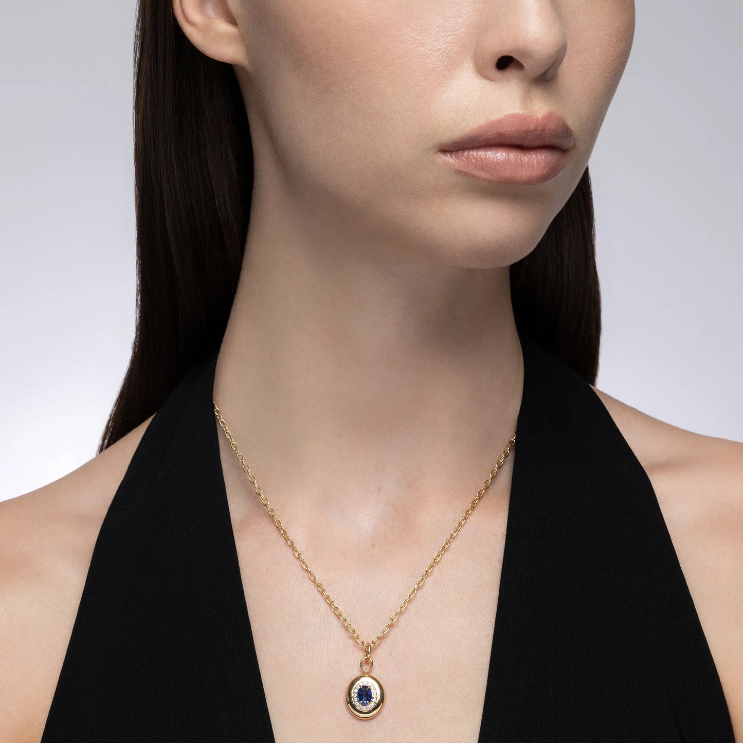A close-up of a model wearing a delicate gold chain necklace with a circular pendant featuring a deep blue center stone.