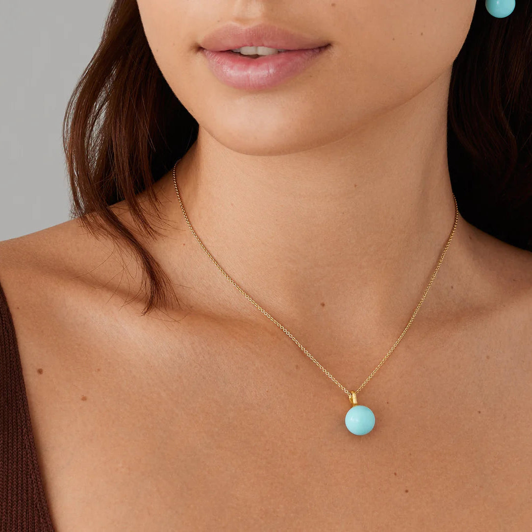 Close-up of a person wearing a gold necklace with a turquoise pendant against a neutral background