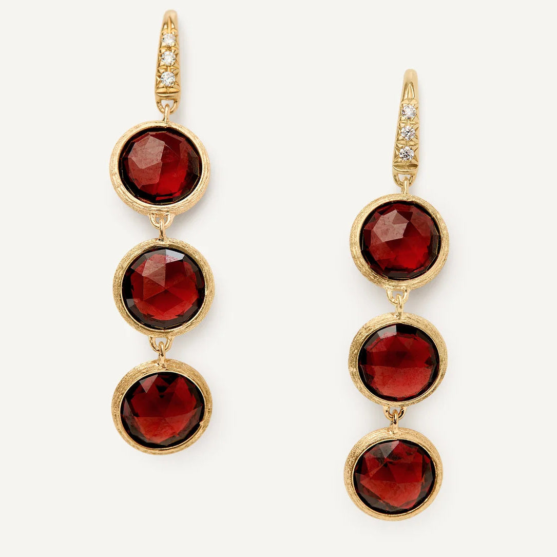 A pair of gold drop earrings featuring three faceted deep red gemstones in a vertical arrangement, with small clear stones along the top hooks, set against a plain off-white background.