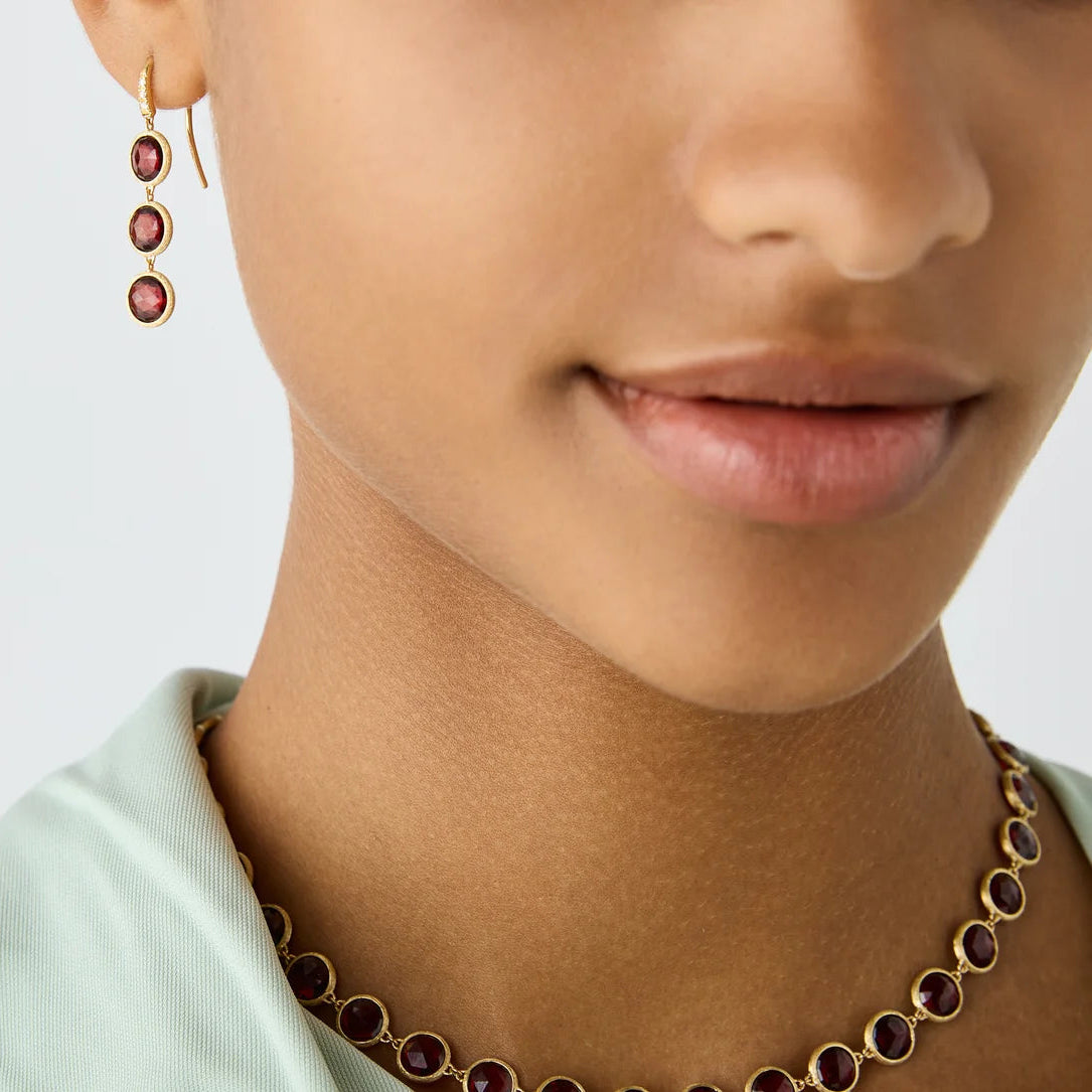 Close-up of a model wearing a gold necklace and matching drop earrings with round red garnet gemstones, set against a clean light gray background.