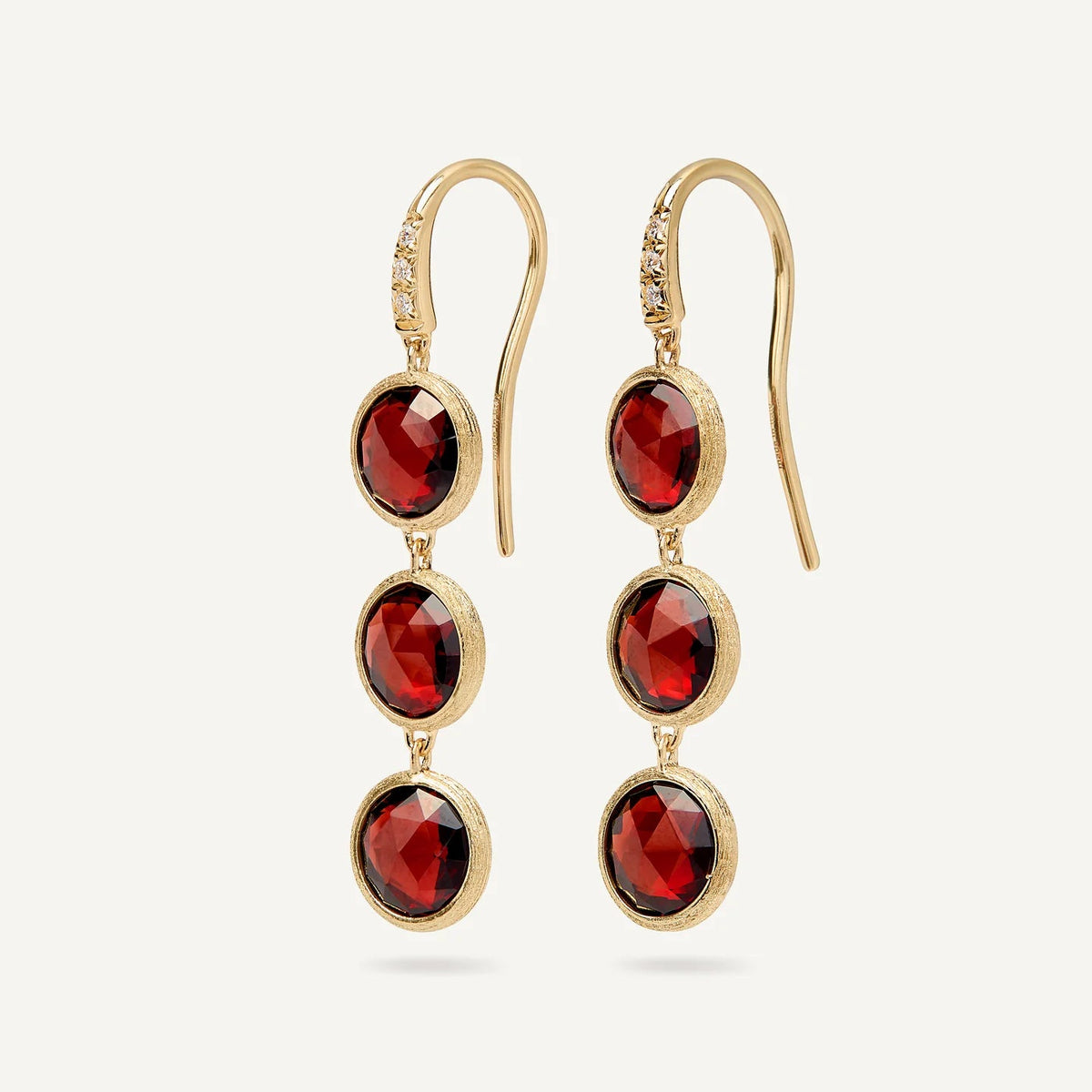 A pair of gold drop earrings featuring three faceted round red garnet gemstones in a textured gold bezel setting, suspended from gold French hooks with a small clear accent stone near the top.