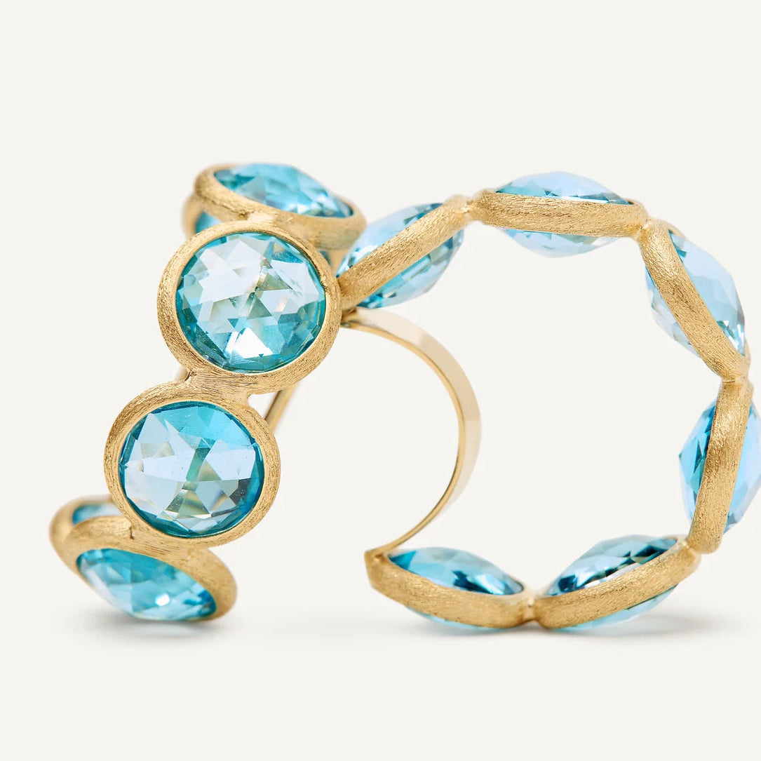 A yellow gold hand engraved hoop featuring large, faceted blue topaz gemstones set in a textured gold bezel setting, photographed on a clean off-white background.