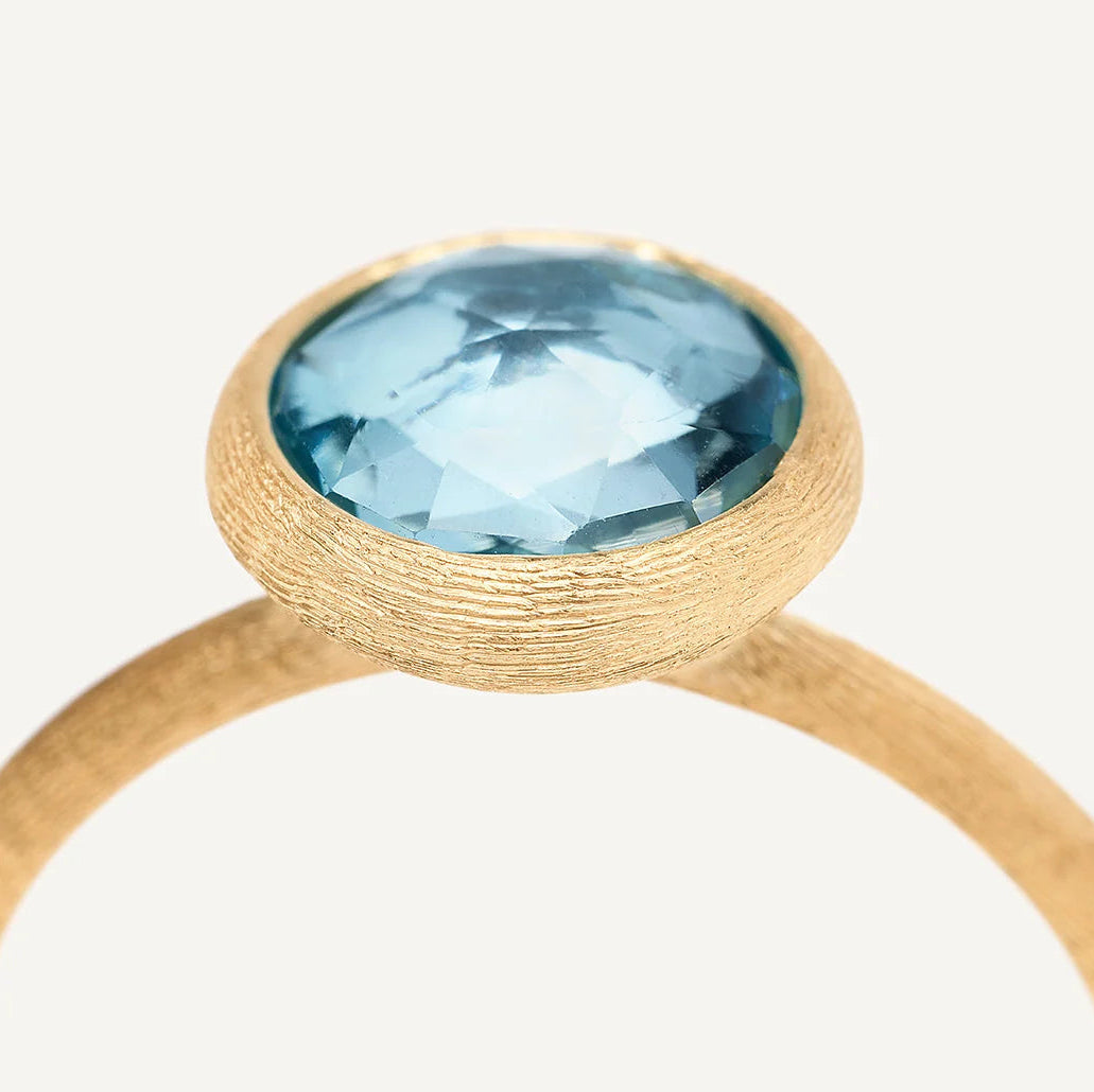 A close-up shot of a gold ring featuring a round, hand-faceted blue topaz gemstone in a textured bezel setting, shown against a plain off-white background.