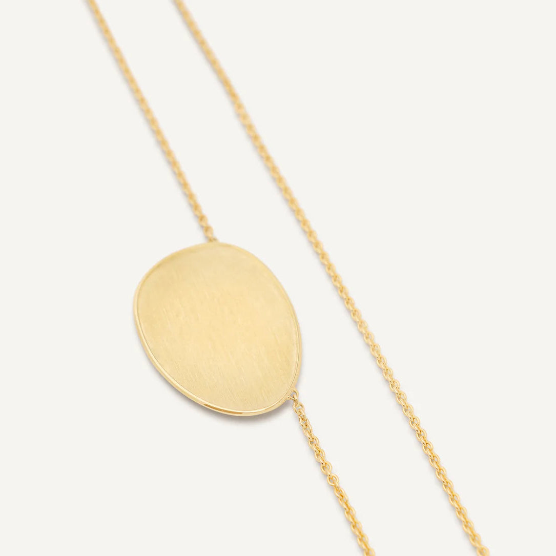 A delicate gold chain necklace featuring a large, flat, oval disc pendant with a brushed texture, displayed on a clean off-white background.